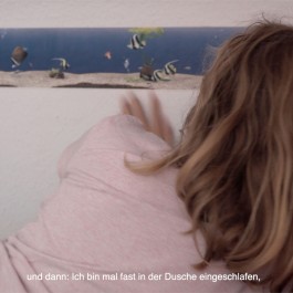 Ausschnitt eines Filmstills des Essayfilms Ich teile mein Bett mit dir. Teil des Kopfes und die linke Schulter eines auf der Seite liegenden Mädchens sind von hinten zu sehen. Das Mädchen schaut auf die Wand vor ihr. Auf Augenhöhe ist daran ein langer Bildstreifen angebracht, der einen sandigen Meeresboden, bunte Fischchen, eine Wasserpflanze und blaues Wasser zeigt. Darunter steht: Und dann: Ich bin mal fast in der Dusche eingeschlafen. 