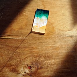 Bildauszug aus der Rauminstallation Mein Fleck der hat drei Ecken: Ein grob geschliffenes Brett, ein Streifen Sonnenlicht fällt diagonal von links unten nach rechts oben darüber. Das Brett hat Risse, ein Astloch ist zu sehen, hier ist ein Stück Holz herausgebrochen. Links oben liegt noch etwas Sägemehl auf dem Brett. Mittig oben liegt eine etwa Handygroßes farbig marmoriertes Glasrechteck. Die Farben darauf erinnern an eine Welle, die über einen Sandstrand spült. 