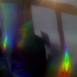 Bild zum Zuhause-Theater 52 Hertz: Das Quadrat zeigt eine spiegelglatte, leicht gewellte, dunkle Fläche, in der sich ein Fensterrahmen spiegelt. Außerdem gibt es links und rechts zwei längliche Streifen, wo sich gleißendes Licht in allen Regenbogenfarben bricht. Von oben über dunkelblau, türkis, grün nach unten zu gelb, orange und rot. Ein Wiederhall des Lichtspiels breitet sich von den beiden Stellen über das ganze Quadrat aus und sammelt sich nochmal in einer Wellenlinie in der Mitte des Quadrats. 