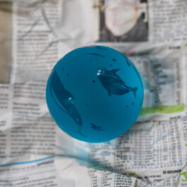 Ausschnitt einer Fotografie der Werkreihe Das ist (ja) voll mein Ding: Den Hintergrund bildet, leicht verschwommen, eine französische Tageszeitung. Darauf, prominent in der Mitte des Bildes, liegt eine türkis, oder besser gesagt meeresfarbene Glaskugel. In die Kugel sind die Formen von Delfinen eingraviert, die als dunkle Schatten sichtbar sind. 
