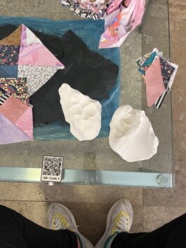 3 - D Tastmodelle, auf einem Glastisch, PatchworkQuadrate zusammengenäht zum Anfassen, Einzelteile aus Stoff, die das Tangrampuzzlquadrat bilden bilden, zum Anfassen, einer der3 QR Codes die zur Audiodeskription führen 