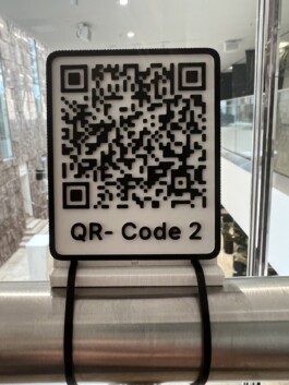 Bild des 3d Print eines der 3 QR Codes, die zur Audiodeskription führen, die die Ausstellung beschreiben. Montiert auf dem Geländer. Am Anfang der Ausstellung. 